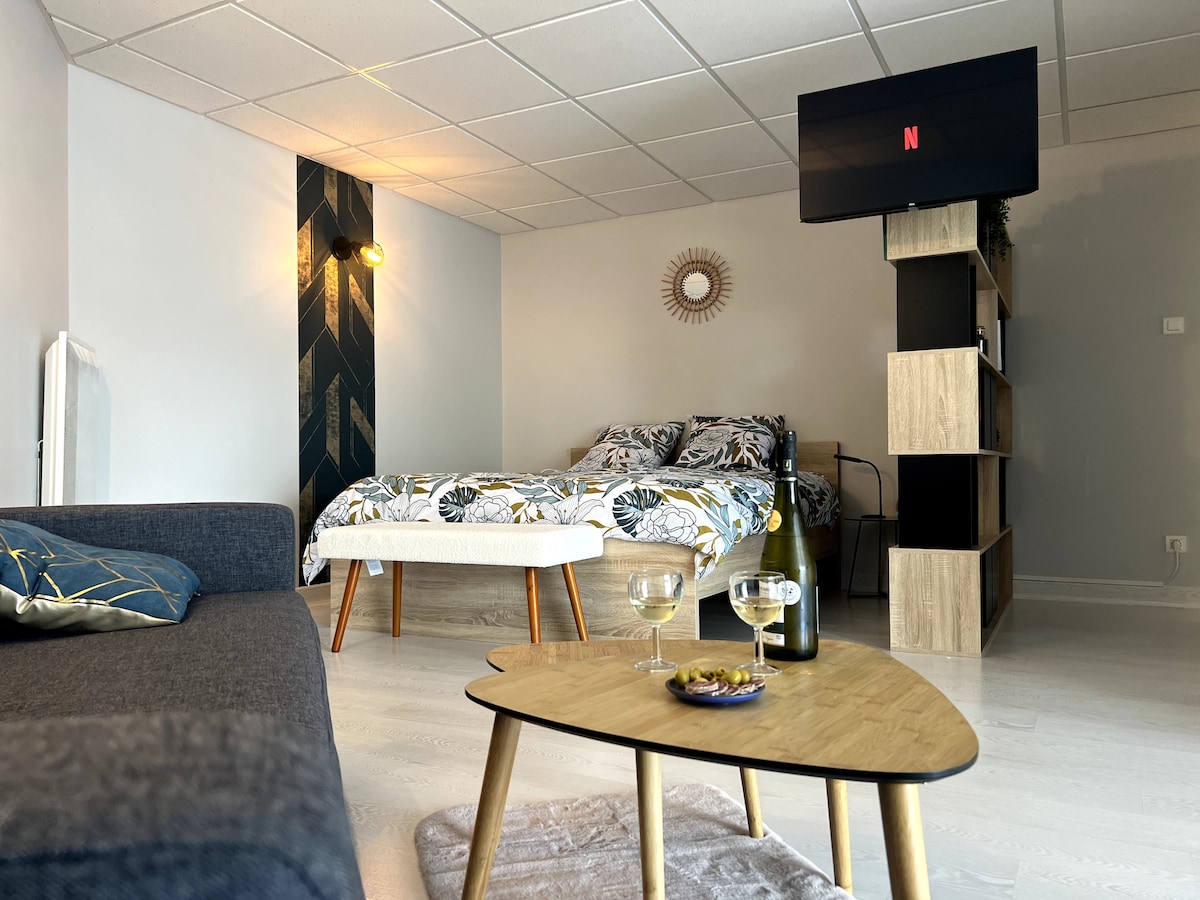 An open living space features a cozy sofa and a low wooden coffee table, which holds two glasses and a bottle. A spacious bed with a floral-patterned duvet is visible, and a wall-mounted television provides entertainment options. Natural light fills the area.