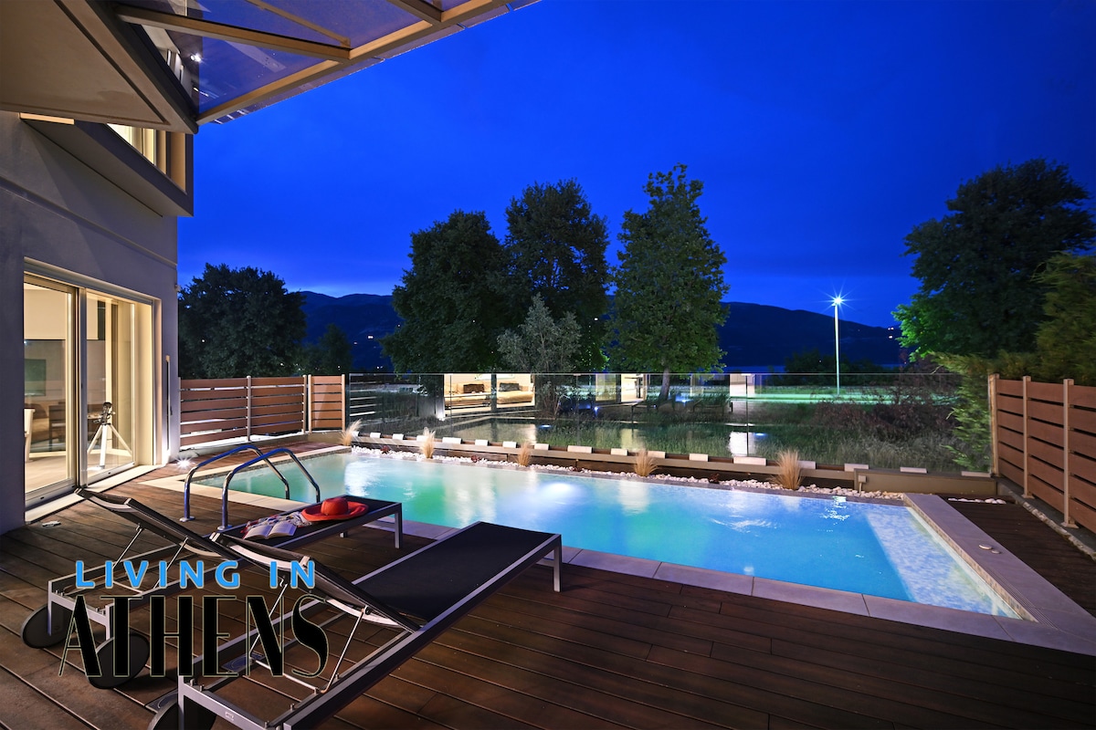 A modern outdoor area is highlighted by a pool reflecting the evening sky. Lounge chairs are positioned nearby on a wooden deck. Lush trees and a serene landscape are visible in the background, contributing to a peaceful ambiance.