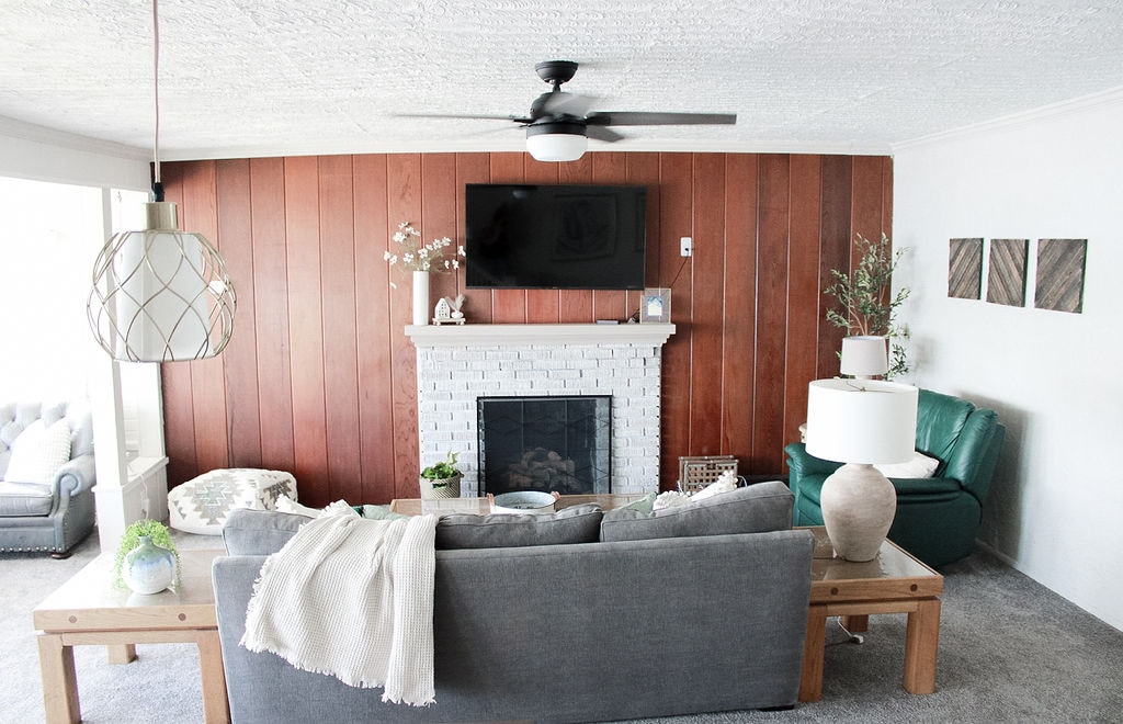A spacious living room is featured, showcasing a cozy sofa and a wooden coffee table. A fireplace acts as a focal point, topped with a flat-screen TV. Natural light is let in through large windows, enhancing the warm wood paneling and soft furnishings.