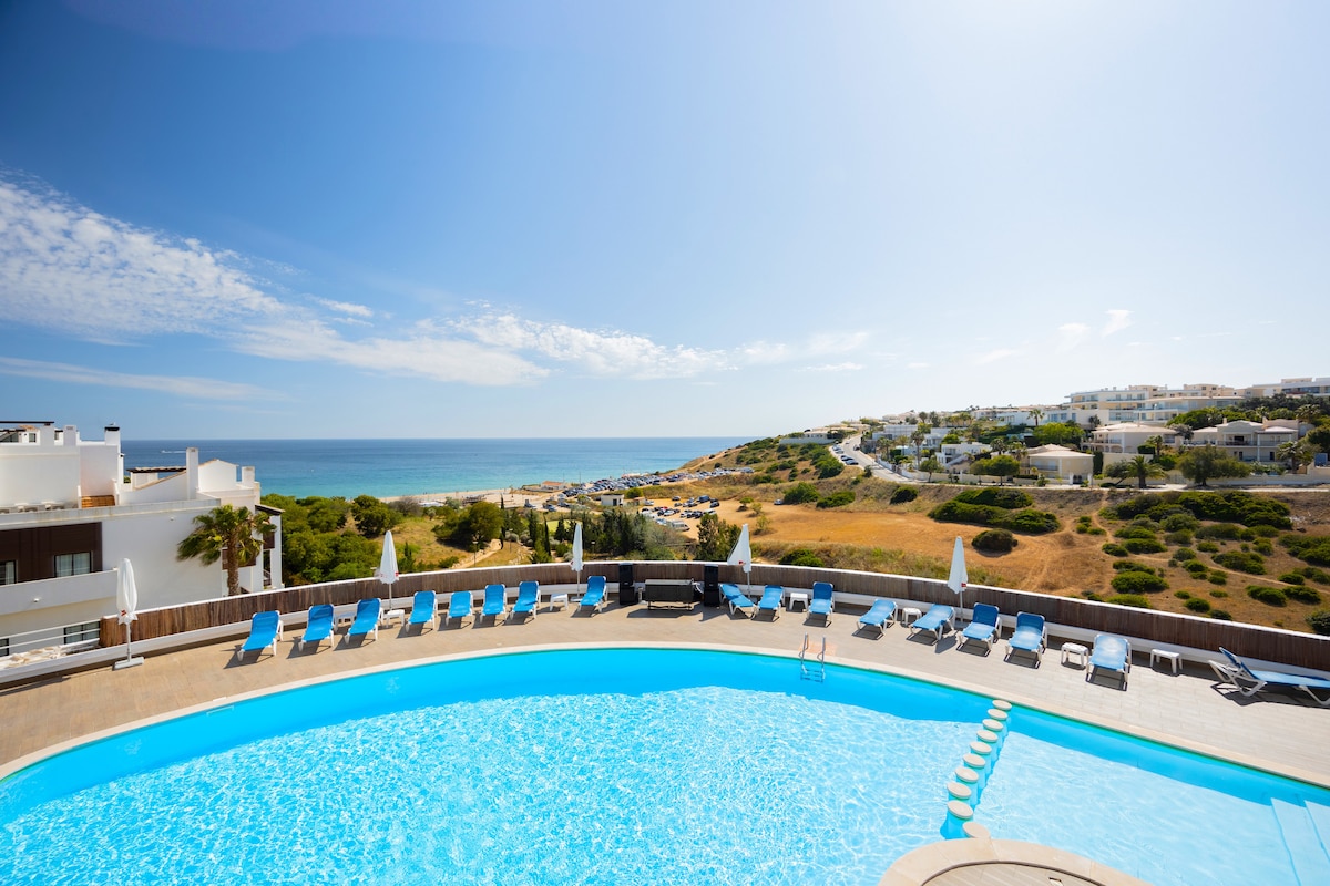 A large outdoor pool is featured, surrounded by sun loungers and umbrellas. Clear blue water reflects the sunlight, while a scenic view of the coastline and rolling hills is visible in the background, enhancing the relaxing atmosphere of the area.