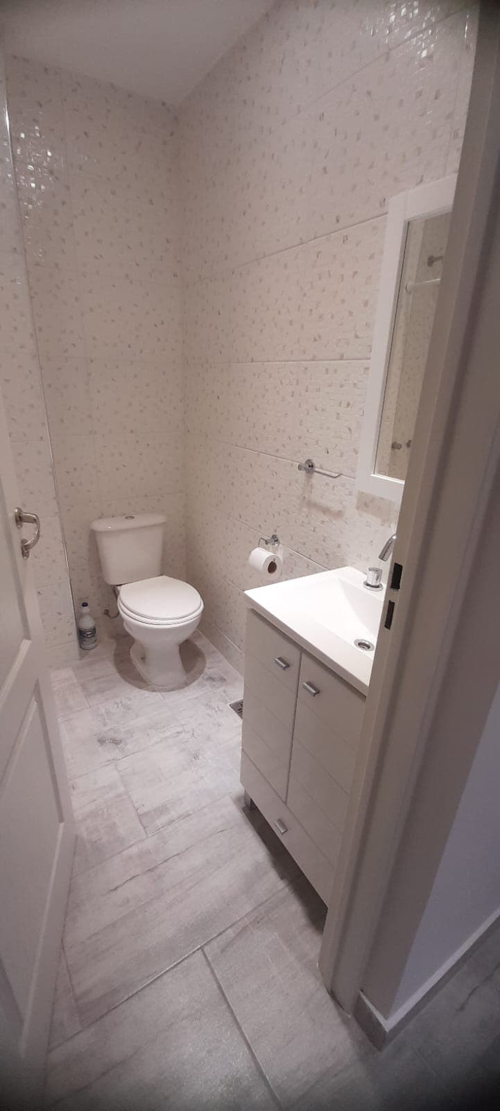 A well-lit bathroom features neutral-colored walls adorned with a subtle floral pattern. A clean, white toilet is positioned beside a small vanity with a sink and a mirror above it. Light-colored tiled flooring enhances the space's brightness.