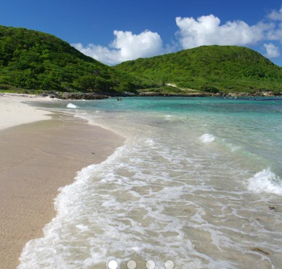 A serene beach scene showcases soft, sandy shores meeting gentle turquoise waves. Lush green hills rise in the background, framed by a clear blue sky dotted with fluffy white clouds.