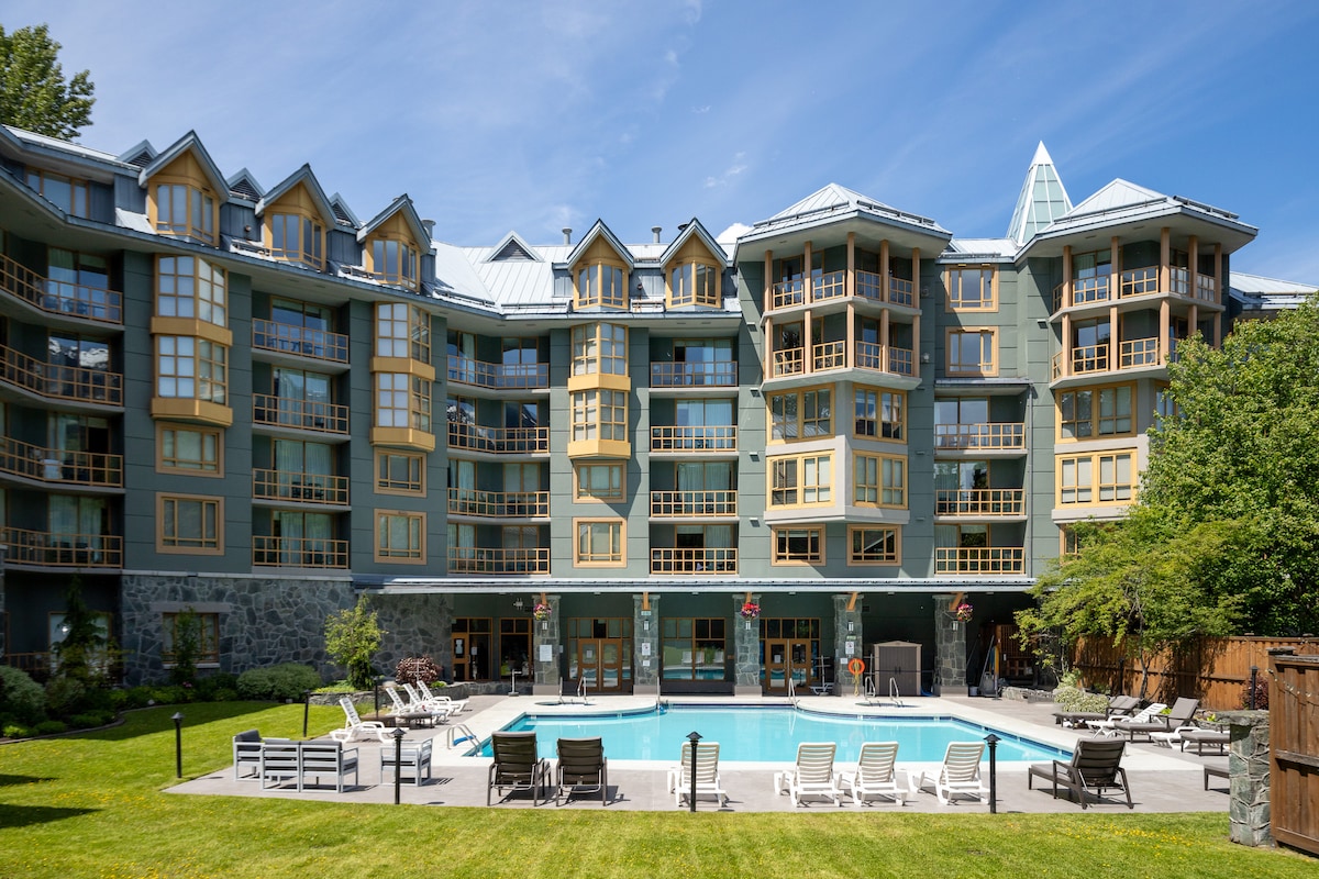 The exterior of a multi-story building is displayed, featuring a mix of balcony designs and large windows. A swimming pool is centrally located, surrounded by lounge chairs and manicured green spaces, creating an inviting atmosphere for relaxation.