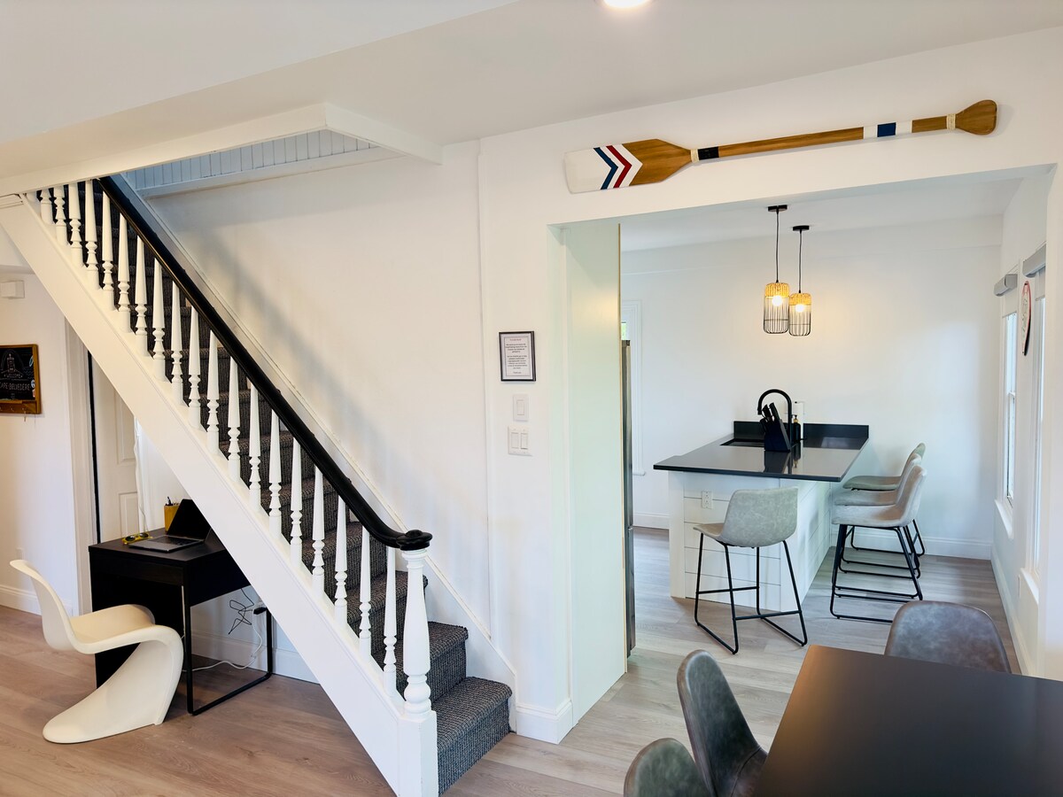 The open living space features a staircase leading to the upper level, accompanied by sleek white walls and wood accents. A decorative paddle hangs above the entry to the modern kitchen, which includes a black island and four bar stools.