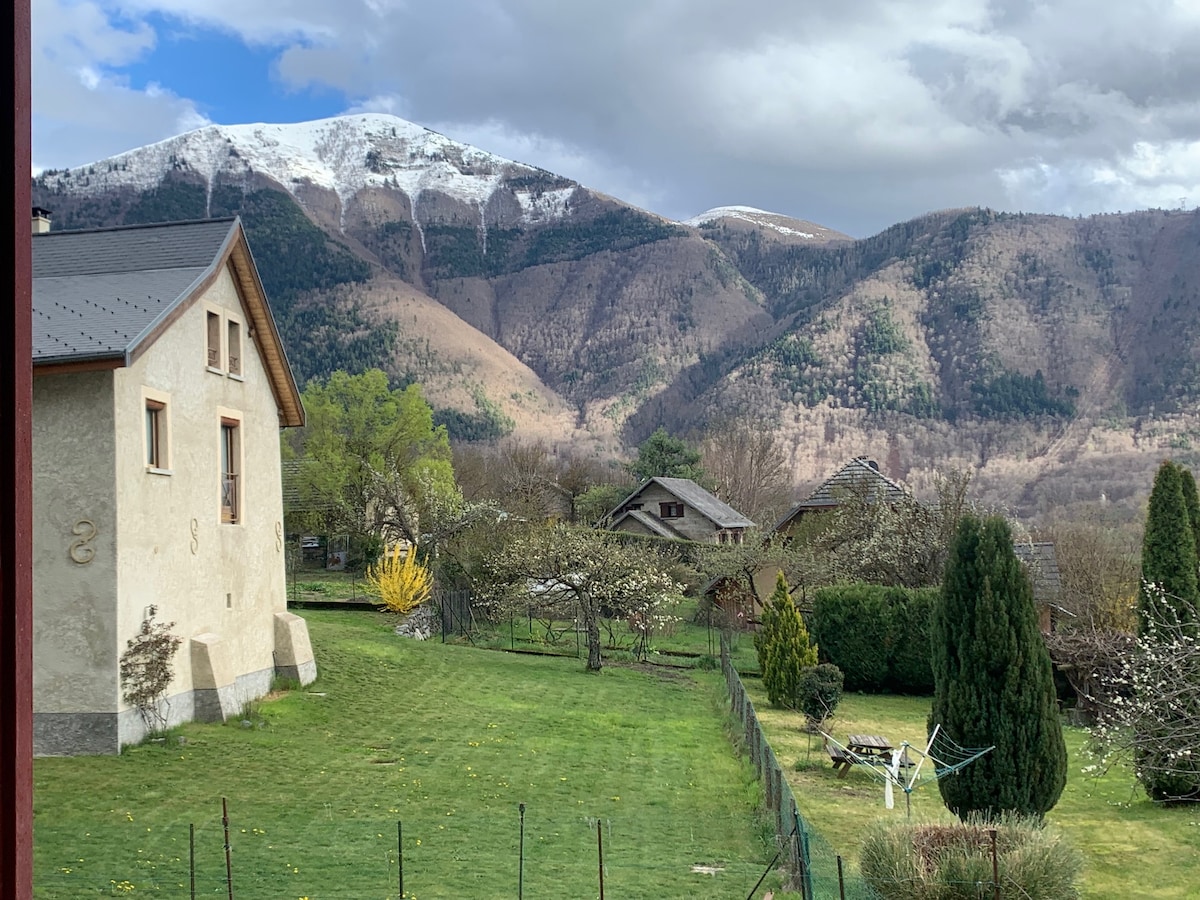A serene outdoor view features a well-kept lawn framed by trees and a small garden area. Snow-capped mountains rise majestically in the background, while several houses dot the landscape, creating a peaceful rural atmosphere.