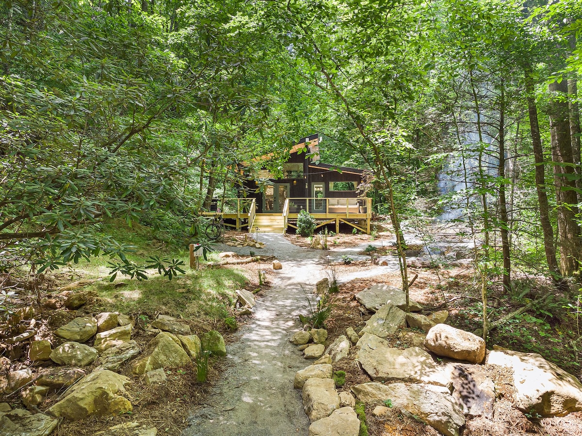 A modern cabin nestled among lush greenery, showcasing a pathway lined with stones leading to the entrance. The structure features a large, inviting deck and is surrounded by tall trees, creating a serene atmosphere in nature.