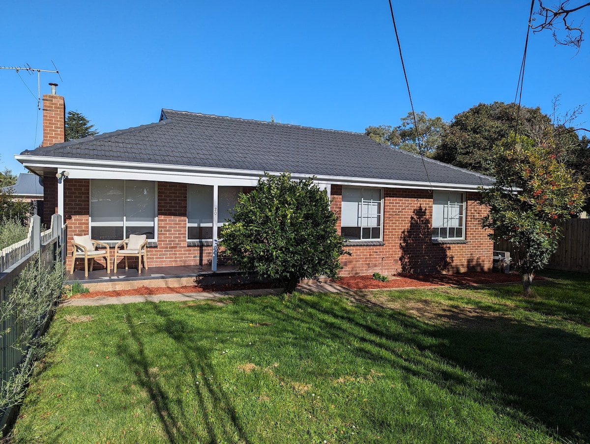 A single-story cottage features a brick exterior with a dark roof. Two seating chairs are placed on a patio in front of large windows, providing views of the well-maintained lawn and surrounding greenery. The space offers a sense of openness and tranquility.