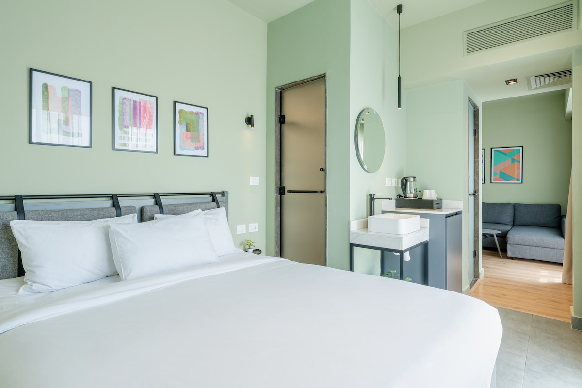 A light-filled studio features a spacious bed positioned against vibrant green walls, surrounded by framed artwork. A comfortable seating area and kitchenette are visible in the background, contributing to a modern and functional living space.