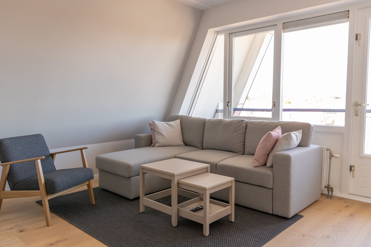 A comfortable seating area features a light gray sectional sofa adorned with pink cushions. A minimalist coffee table is positioned in front of the sofa, while a small accent chair enhances the space. Large windows allow natural light to illuminate the room.