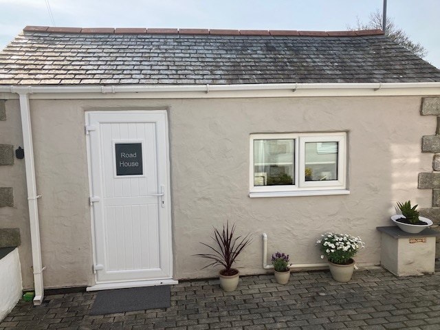 The exterior of the converted garage is shown, featuring smooth, pale stone walls and a sloped slate roof. A white door marked 'Road House' stands at the center, flanked by two windows. Potted plants are arranged along the entrance, enhancing the welcoming appearance.