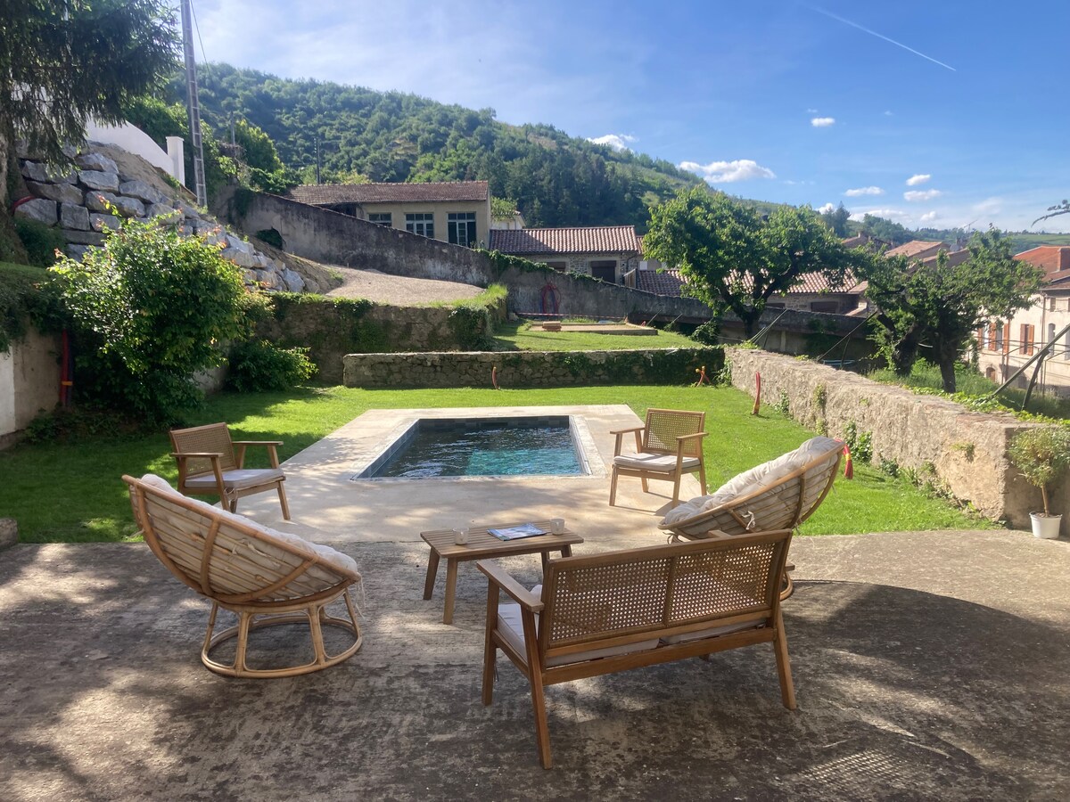 A serene outdoor area is captured with a swimming pool at the center, surrounded by lush greenery. Comfortable seating is arranged nearby, including wicker chairs on a stone patio. The backdrop features hills and buildings, offering a peaceful ambiance.