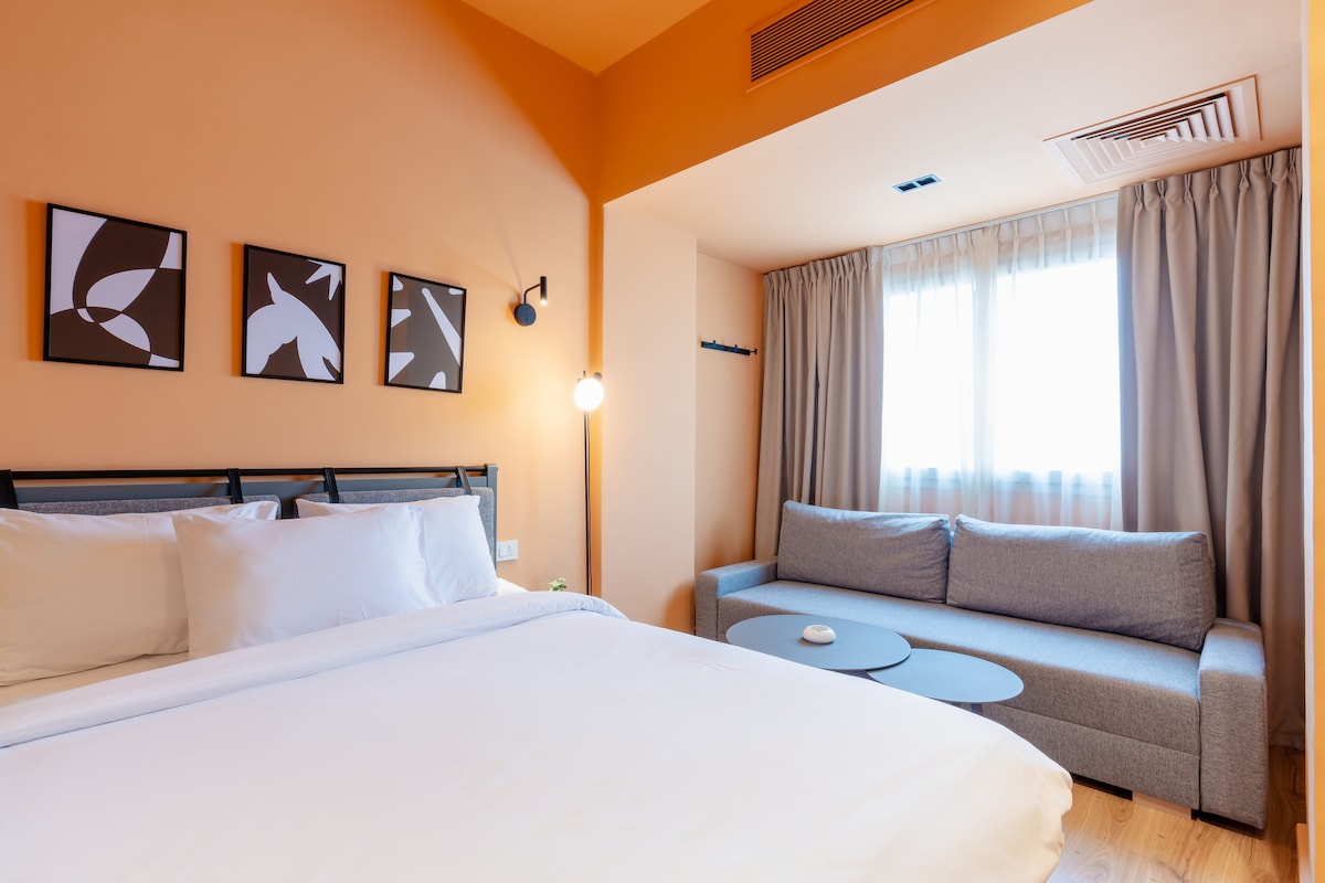 A modern studio features a spacious bed with crisp white linens, complemented by framed abstract art on the walls. A comfortable grey sofa is positioned near a sleek coffee table, and natural light filters through a large window covered with light curtains.