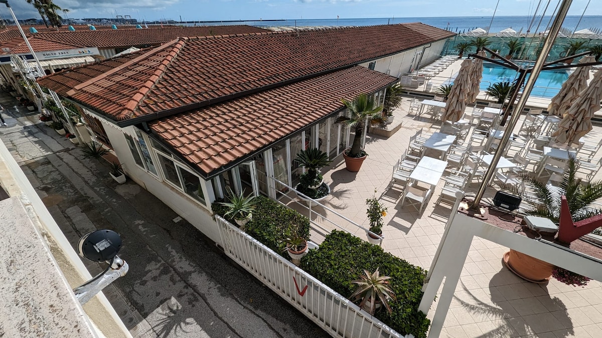 The image displays a view of a coastal establishment with a tiled roof and several outdoor seating areas. Sun loungers and shaded umbrellas are visible, surrounded by landscaped greenery, while the tranquil sea can be seen in the background under a partly cloudy sky.