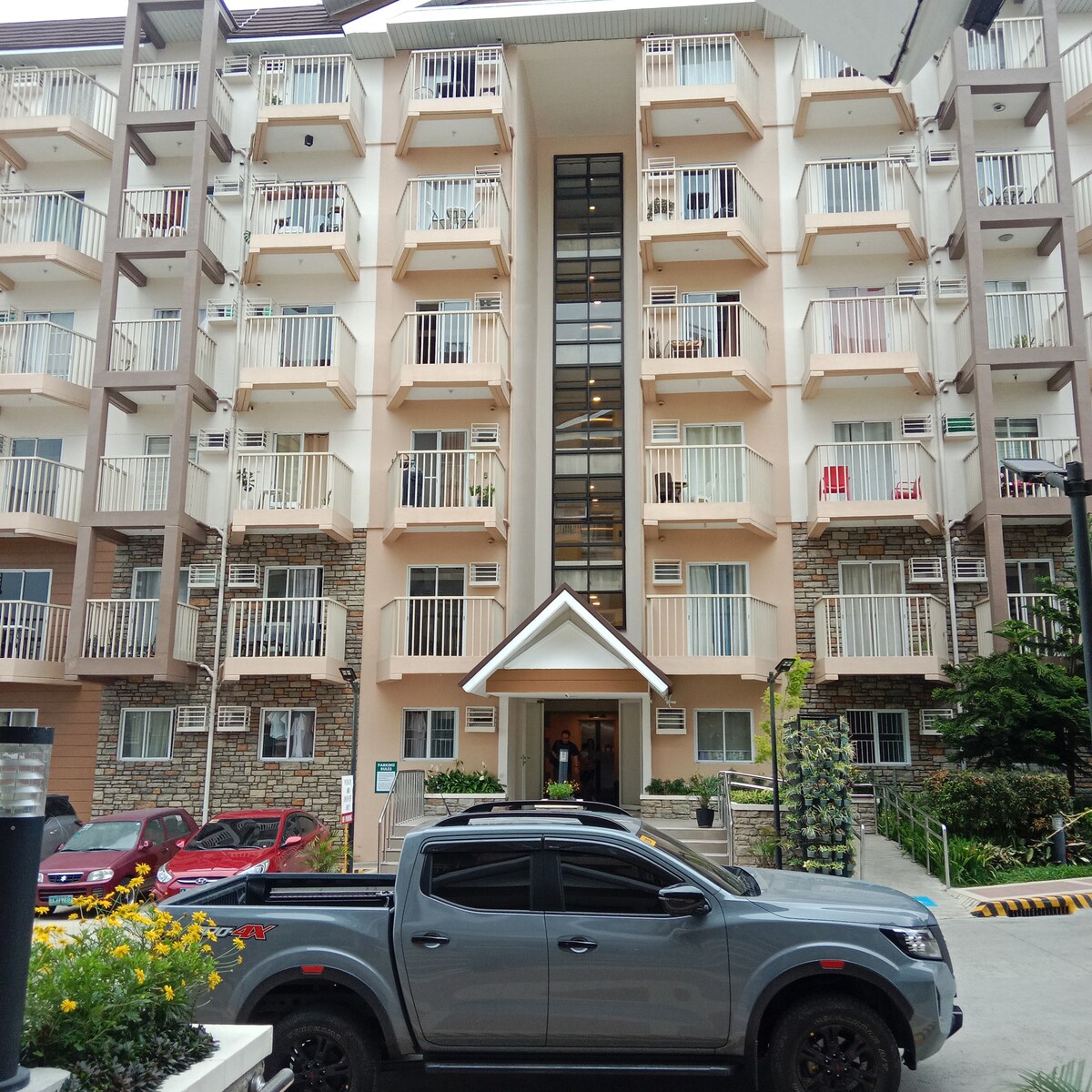 The exterior view of a multi-story condominium building is presented, showcasing a modern design with balconies on each level. A welcoming entrance is visible at the center, framed by greenery. A parked grey vehicle is positioned in the foreground, indicating available parking.