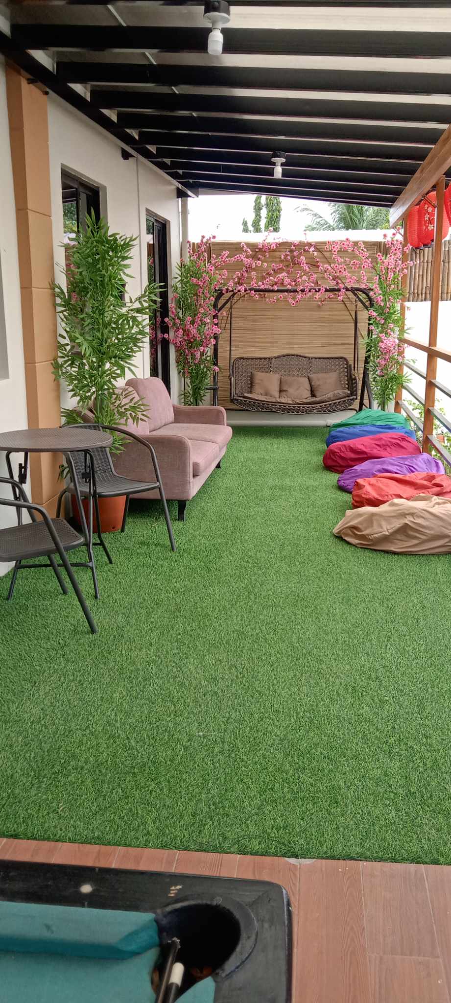 An outdoor patio area features green artificial grass and is decorated with potted plants and pink flowering vines. A cozy swing chair and colorful beanbags offer comfortable seating options, complemented by a small table and two chairs for relaxation.