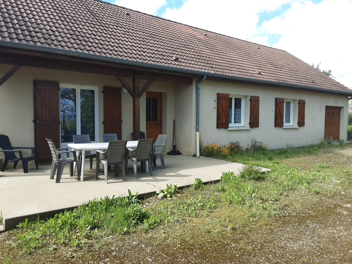 A single-story house is featured, showcasing a covered patio with a large table and several chairs set on a concrete surface. The exterior walls are light-colored, and the windows are adorned with shutters. Lush greenery and wildflowers are visible in the garden area.
