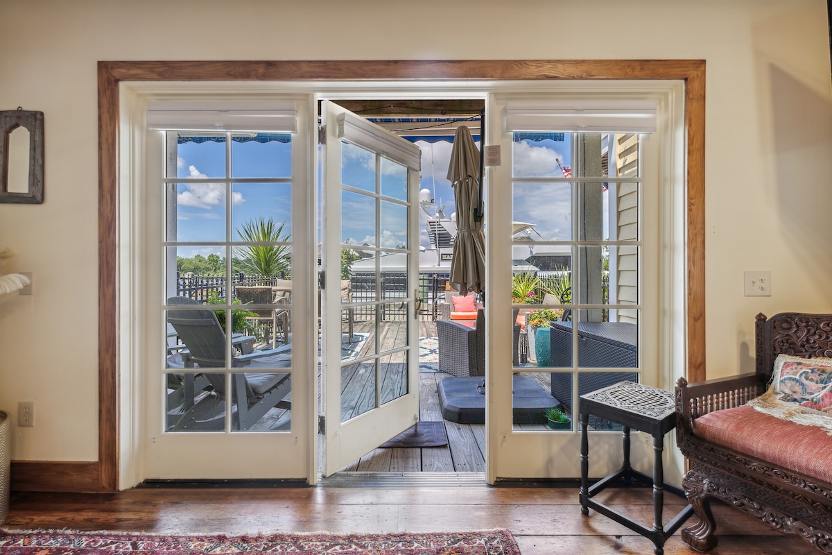 Double doors open to a sunny patio featuring comfortable seating and potted plants. The view includes outdoor furniture arranged for relaxation, with blue skies and a few clouds visible above. Rustic wooden flooring extends into the patio area, creating a cohesive indoor-outdoor feel.