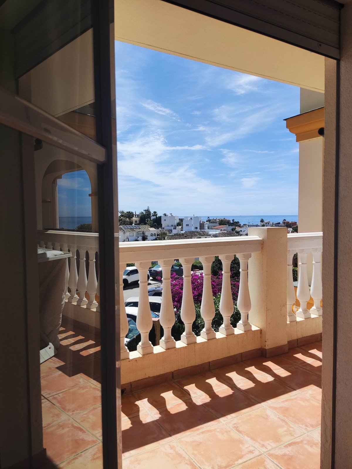 A balcony view is captured, featuring a railing with classic balusters. In the distance, the sea is visible under a clear blue sky, while vibrant pink bougainvillea can be seen framing the scene, enhancing the outdoor space's appeal.