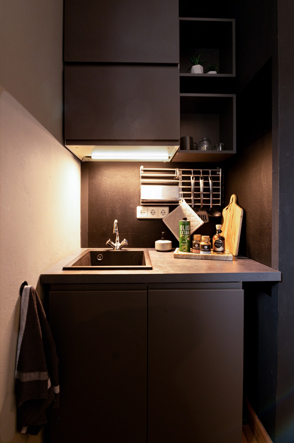 A compact kitchenette is seen featuring a sink, a small countertop, and minimalistic storage above. Kitchen supplies and decorative items are neatly arranged on the shelves. A towel hangs nearby, while ambient lighting enhances the modern design.