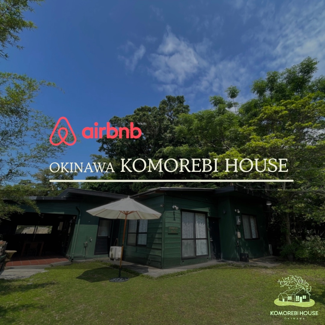A green house is set within a lush garden, featuring a large umbrella over a seating area. Tall trees provide shade, and a spacious lawn is seen in front. The exterior is cheerful, blending harmoniously with the natural surroundings.