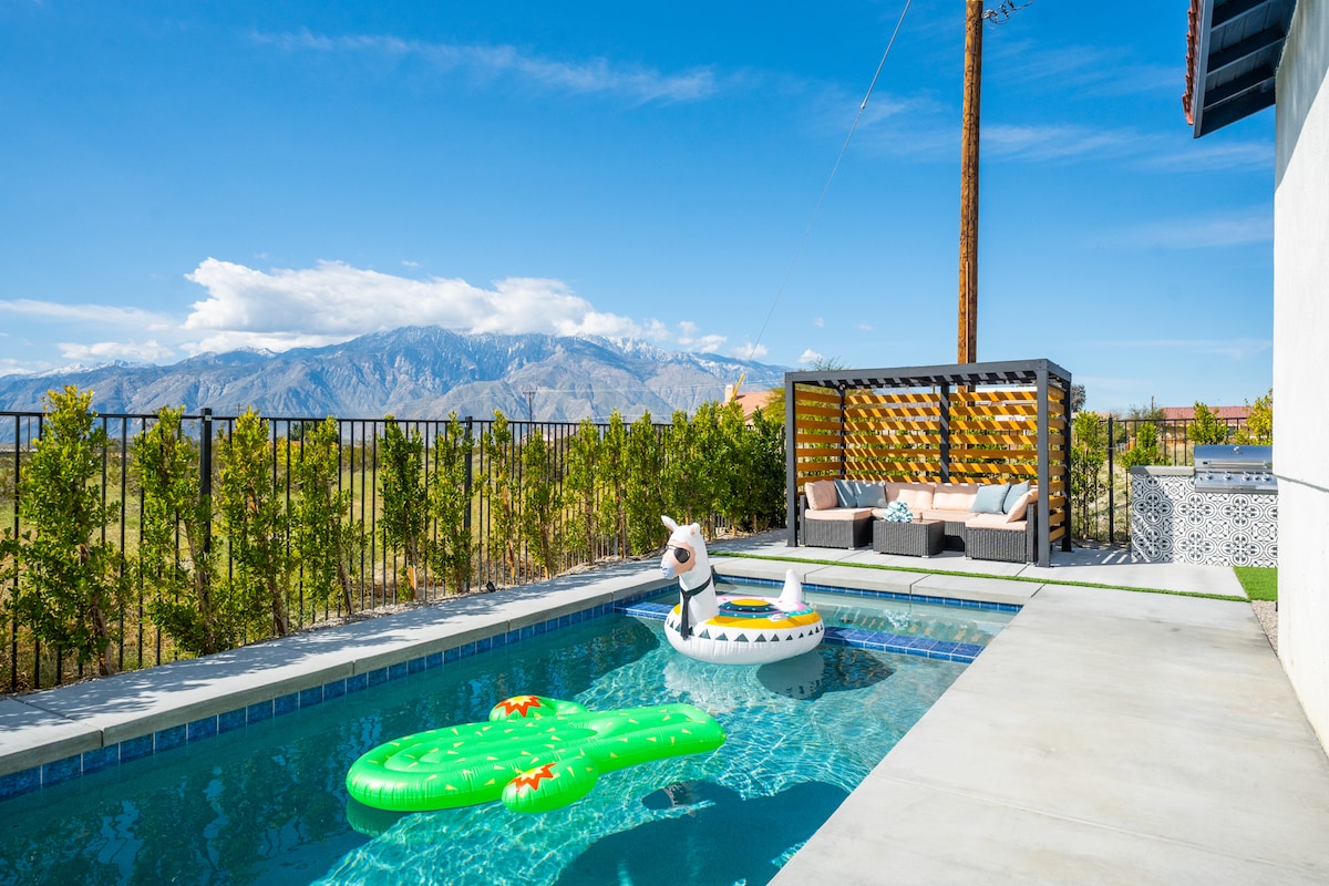 A private pool area features a green inflatable alligator and a white float shaped like a dog, inviting relaxation. Mountains rise in the background under a clear blue sky, while a shaded seating area is positioned beside the pool, offering a space to unwind.