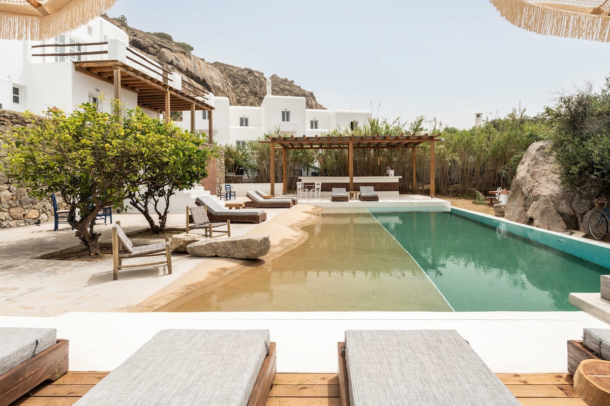 The shared outdoor pool area features a blend of natural elements and modern design. Comfortable lounge chairs are positioned on wooden decking, while a pergola provides shade over a spacious dining area. Lush greenery and smooth stone create a serene environment near the pool.