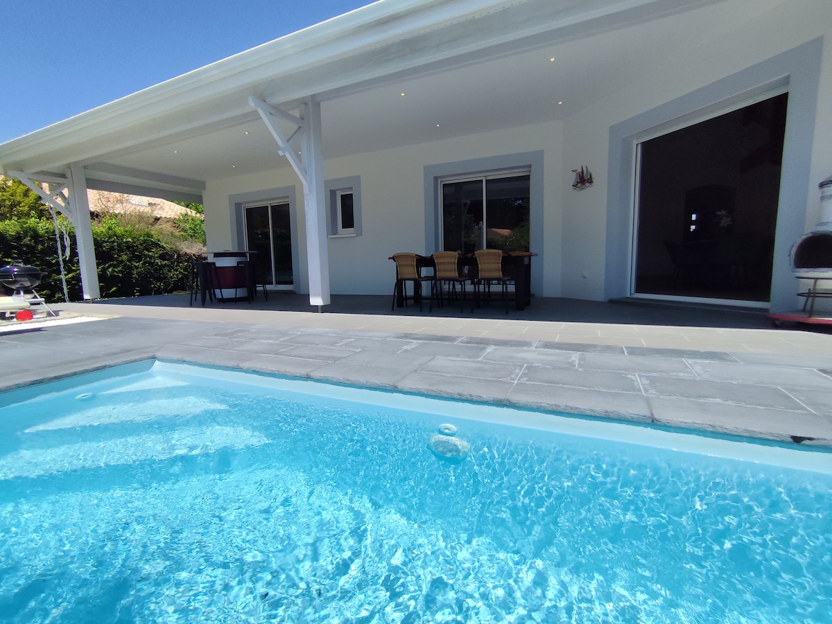 An outdoor area features a crystal-clear private pool in front of a covered terrace. The terrace is furnished with a dining table and chairs, providing a space for relaxation and enjoyment. Large glass doors open to the interior, allowing natural light to enter.