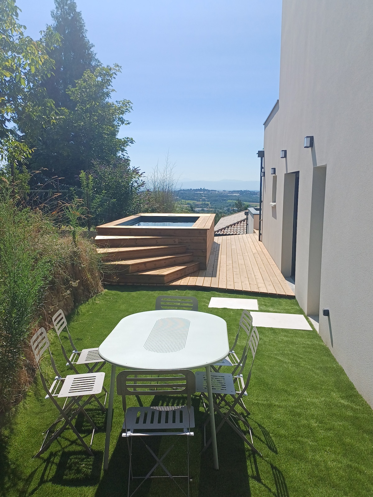 An outdoor area showcases a wooden deck adjacent to a private pool surrounded by greenery. A table with metal chairs is positioned on a well-maintained lawn, offering a serene space for relaxation and dining. The distant landscape provides a scenic backdrop.
