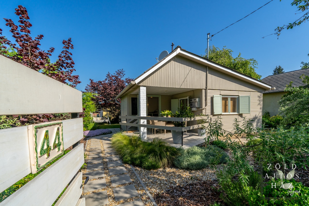 A single-story house with a light exterior is framed by lush green foliage and colorful trees. A covered porch is accessed by a stone pathway lined with gravel, while the house number is prominently displayed on the fence.