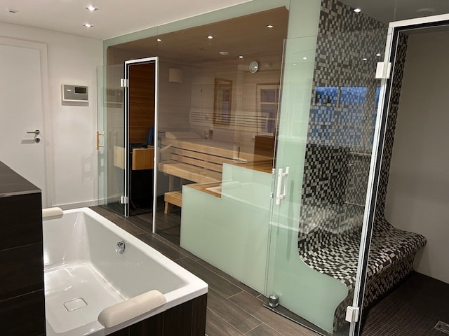 A modern spa area is featured, showcasing a freestanding bathtub in front and a sauna setup behind. The glass walls create a sense of openness, while a relaxation bench and textured seating area offer comfort. Soft lighting enhances the calming atmosphere.
