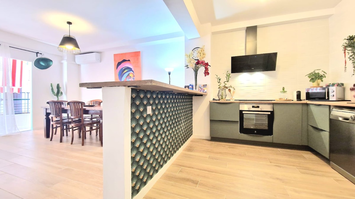 A modern kitchen area features a sleek countertop and is equipped with essential appliances. The dining space is visible with wooden chairs surrounding a large table. Colorful artwork and decorative plants add a vibrant touch to the open layout.