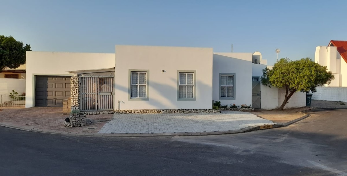 A modern one-story home is highlighted with a clean, white exterior and large windows. A driveway leads to a garage, with a small garden area featuring a well-maintained tree. The house is situated on a corner, providing an unobstructed view of the surrounding area.