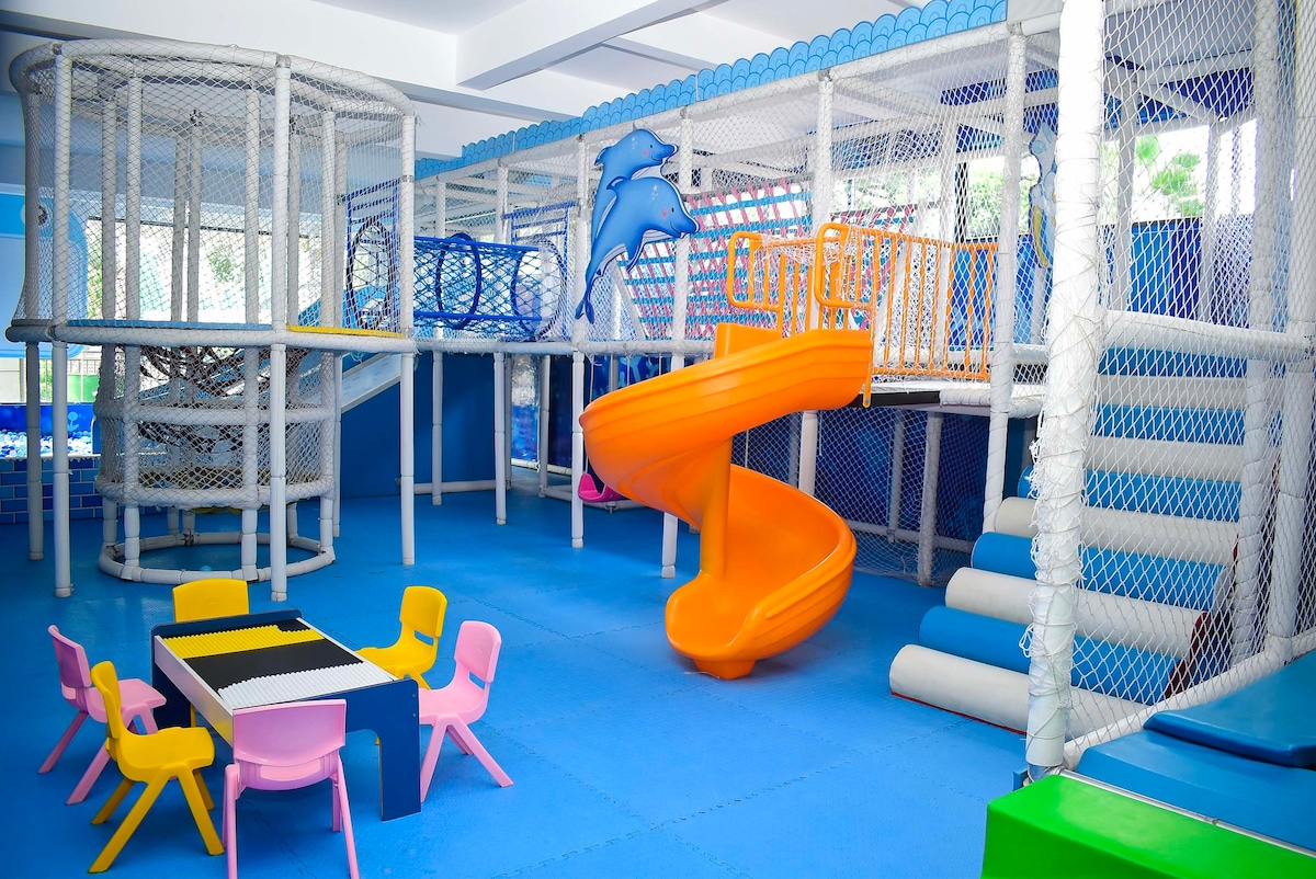 A vibrant children's playground features colorful climbing structures and a large orange slide. Various play areas are enclosed in safety nets. Brightly colored chairs surround a small table, inviting playtime and activities in a spacious, well-lit environment.