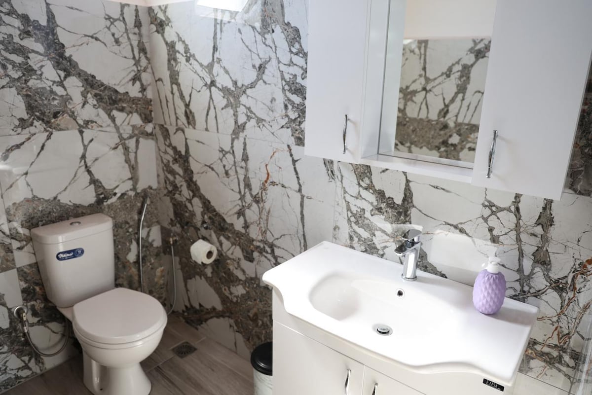 A modern bathroom features striking marble walls with unique veining patterns. A white vanity with a sleek sink is complemented by a large mirror above. A toilet is positioned against the wall, and a decorative lavender item adds a touch of color.