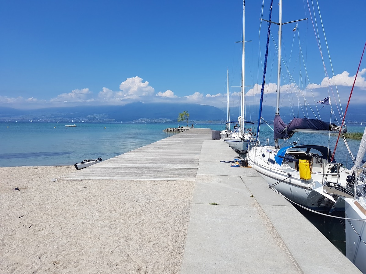 A wooden pier extends into calm waters, flanked by moored sailboats. Soft sand borders the pier, while the backdrop features distant mountains and a clear blue sky with few clouds. Gentle ripples can be seen on the water's surface.