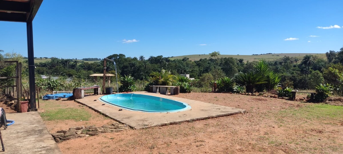 An outdoor area features a rectangular swimming pool, surrounded by a stone patio and greenery. The landscape includes rolling hills, with various plants lining the perimeter. A shaded seating area can be seen in the background, enhancing the relaxing environment.