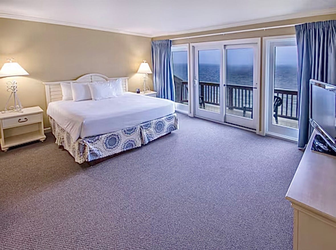 A spacious room features a large bed adorned with white linens. Two bedside lamps are positioned on either side, illuminating the space. Floor-to-ceiling windows offer views overlooking the sea, with a private balcony accessible through sliding glass doors.