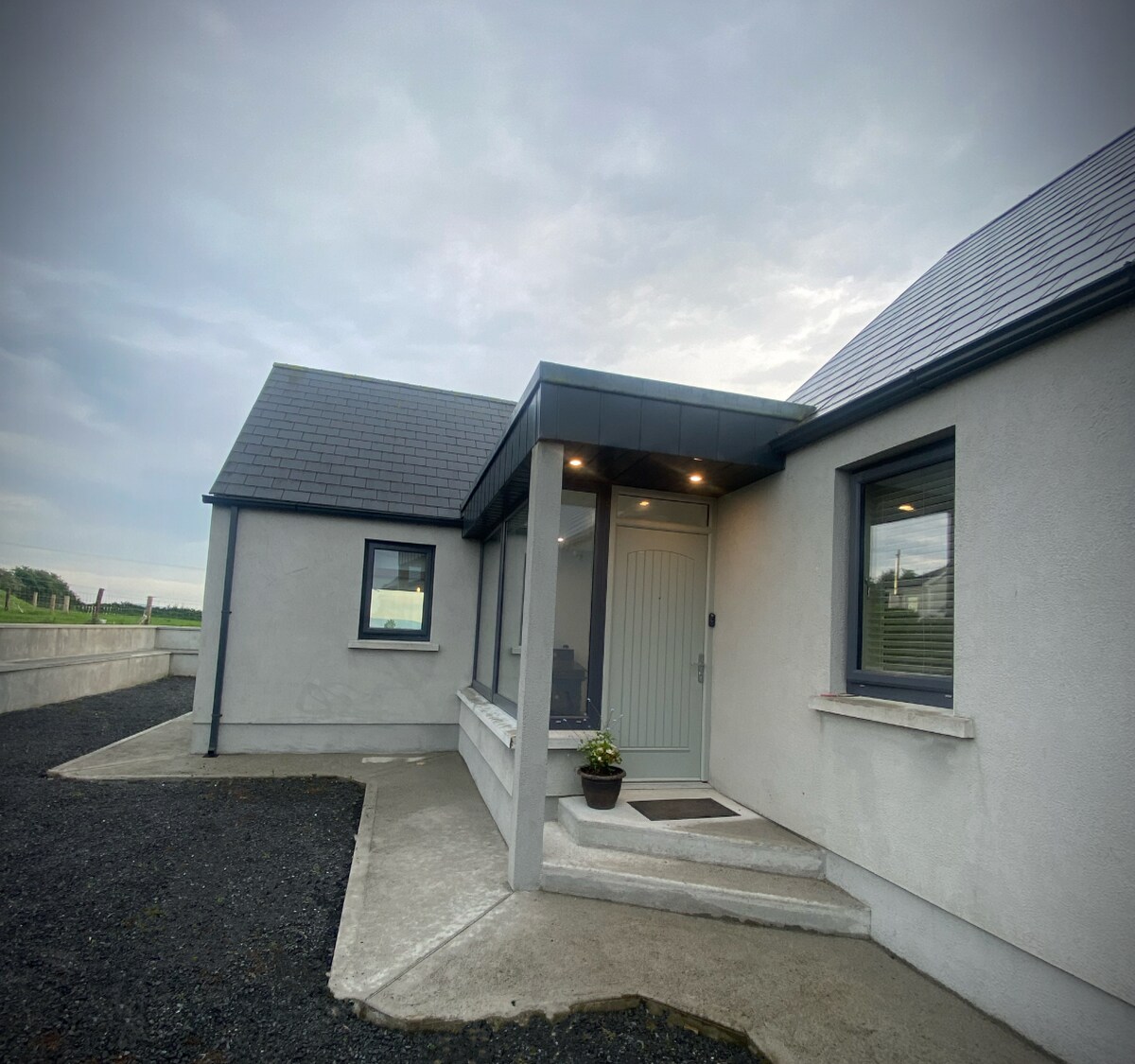 A modern entrance showcases a light-colored door framed by symmetrical windows, leading to a subtle overhang. The exterior walls are finished in light stucco, complementing the gray roof. A small potted plant is positioned on the step, adding a touch of greenery.
