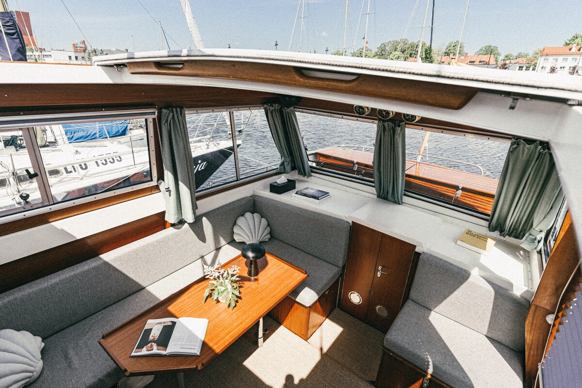 The interior space features comfortable seating arranged around a wooden table. Large windows allow natural light to fill the area, offering views of the water and boats nearby. Soft furnishings in neutral tones complement the classic wood accents.