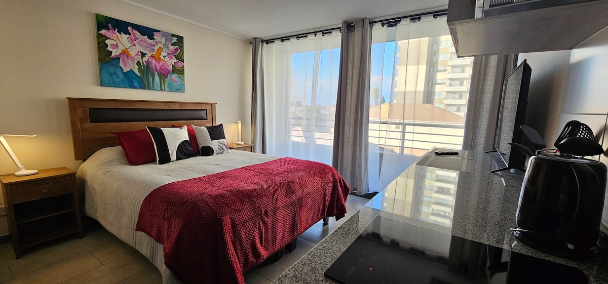 A room featuring a spacious bed dressed in a red and white quilt, complemented by decorative pillows. Natural light filters through sheer curtains, illuminating a small table with an electric kettle and a TV. A colorful artwork enhances the wall above the headboard.