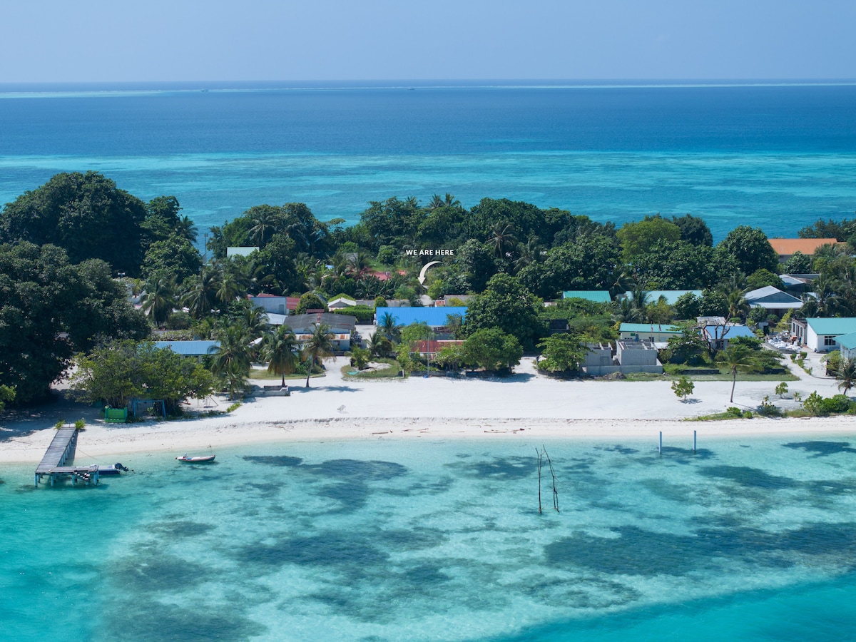 Aerial view of a tranquil island community surrounded by turquoise waters. Sandy shores with scattered palm trees are visible, and a few colorful buildings dot the landscape, blending harmoniously with the natural environment. The clear waters reveal coral reefs and marine life beneath the surface.