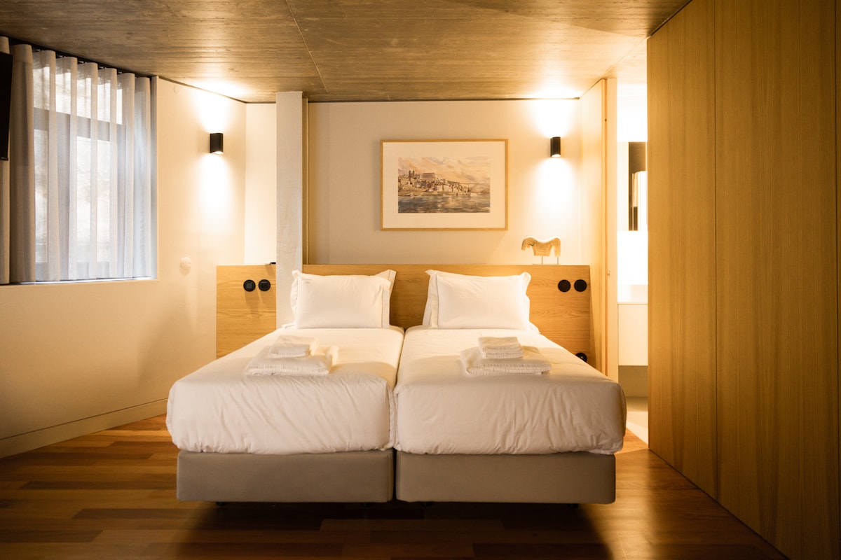 A well-lit room features two spacious beds with clean white linens, arranged side by side. Light wood accents provide a modern touch, while a framed artwork hangs above the beds. Soft curtains allow natural light to filter in from a nearby window.