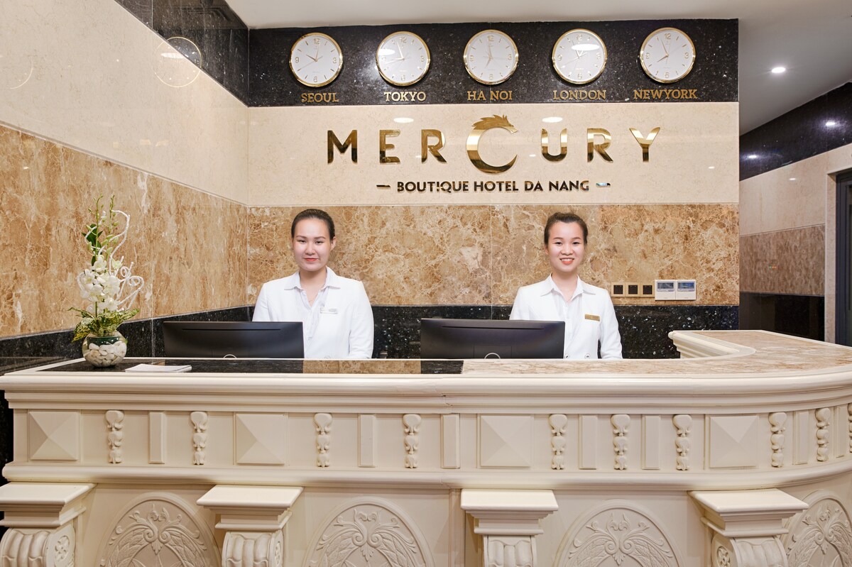 The reception area features a modern design with a marble front desk and elegant wall decor. Clocks displaying different city times adorn the wall above, contributing to a welcoming atmosphere. Two staff members are positioned behind the desk, ready to assist guests.