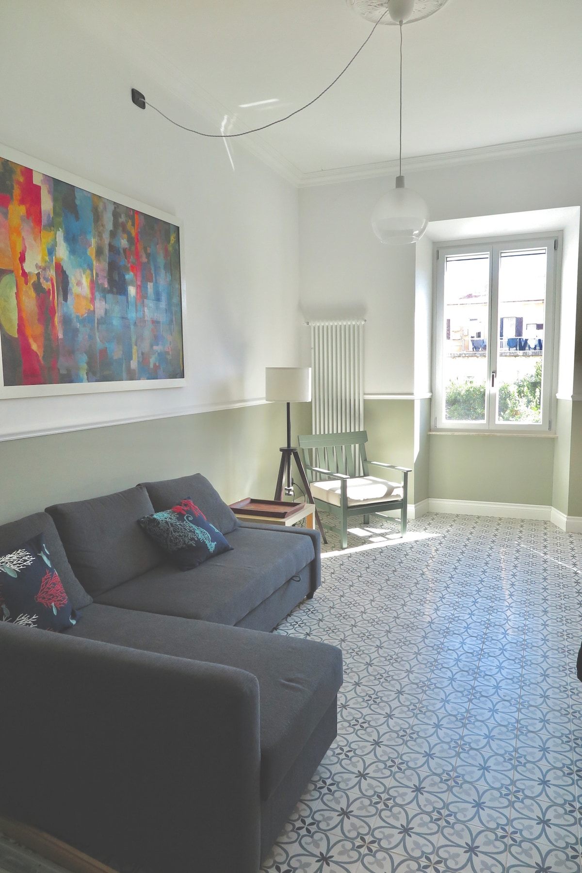 A bright seating area features a gray sofa adorned with colorful cushions. A small green chair sits beside a lamp, and a window allows natural light to fill the space. The floor is decorated with patterned tiles, enhancing the room's elegance.