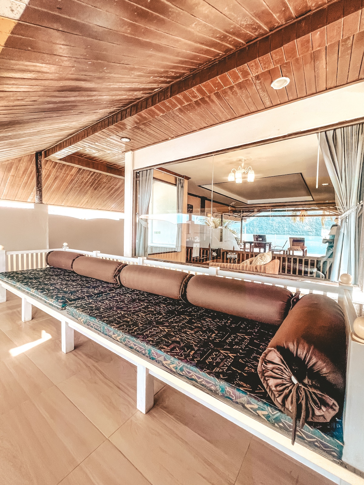 A spacious lounging area is displayed, featuring a long cushioned bench with dark brown pillows. Large windows allow natural light to illuminate the space, and a glimpse of a dining area can be seen in the background, showcasing a calm and inviting environment.