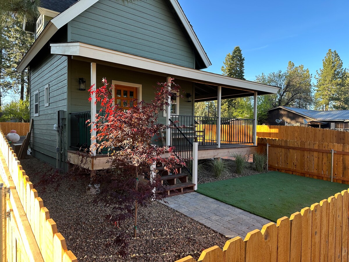 The exterior of a green two-story house is visible, featuring a welcoming front porch with railings. A neatly landscaped area includes a small tree surrounded by gravel and a lawn. A wooden fence encloses the yard, creating a private outdoor space.