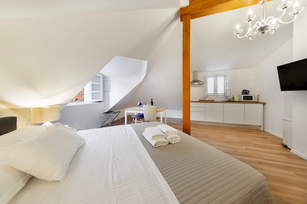 The loft studio features a spacious bed with neatly arranged towels nearby. Natural light fills the room through a window, illuminating the tasteful decor. A dining table and kitchenette are visible in the background, complementing the open layout of the space.