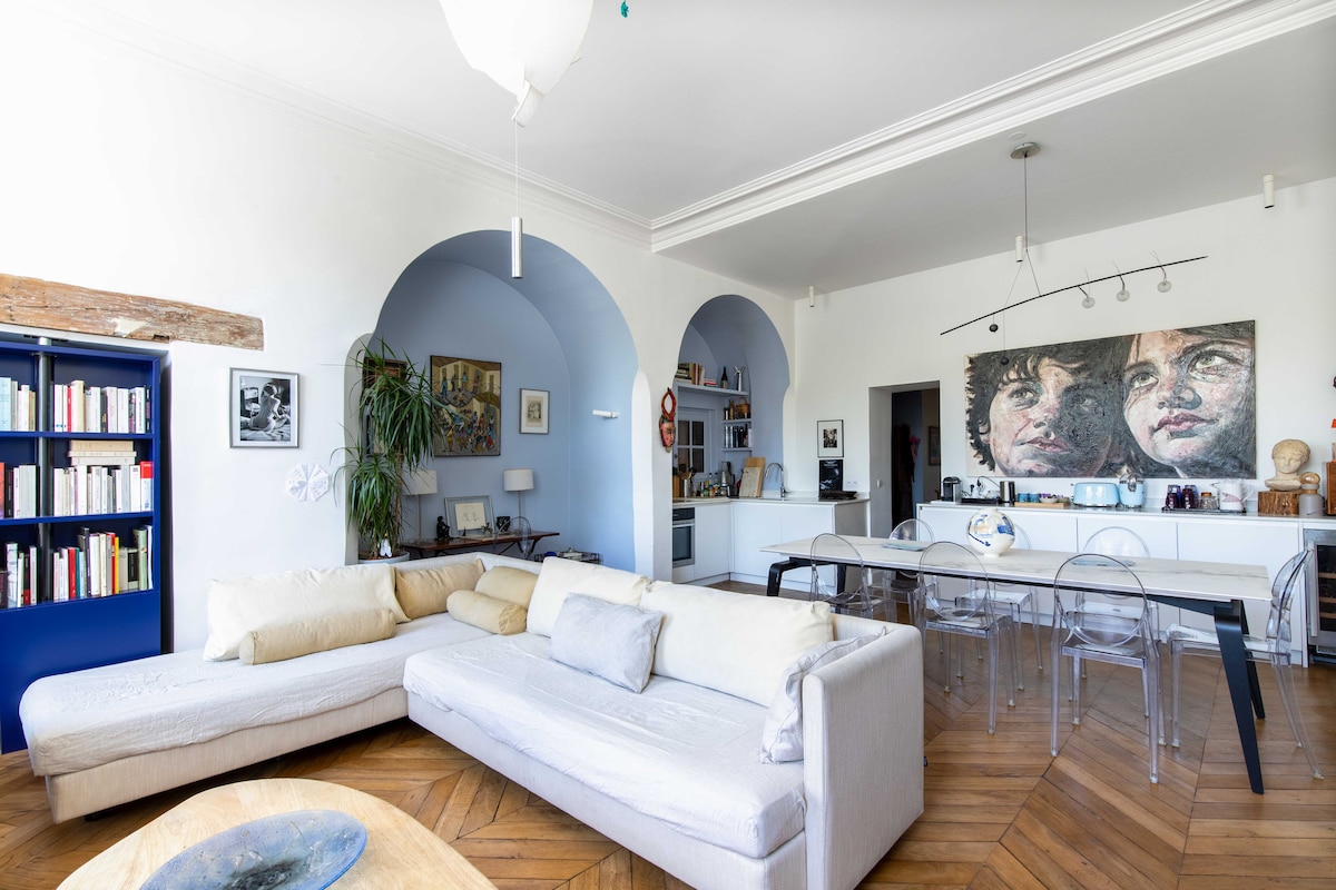 The living area is characterized by a spacious sectional sofa and a light wood coffee table. A dining space with a modern table and clear chairs is positioned near an open kitchen. Shelves filled with books are visible along the wall, adding character to the room.
