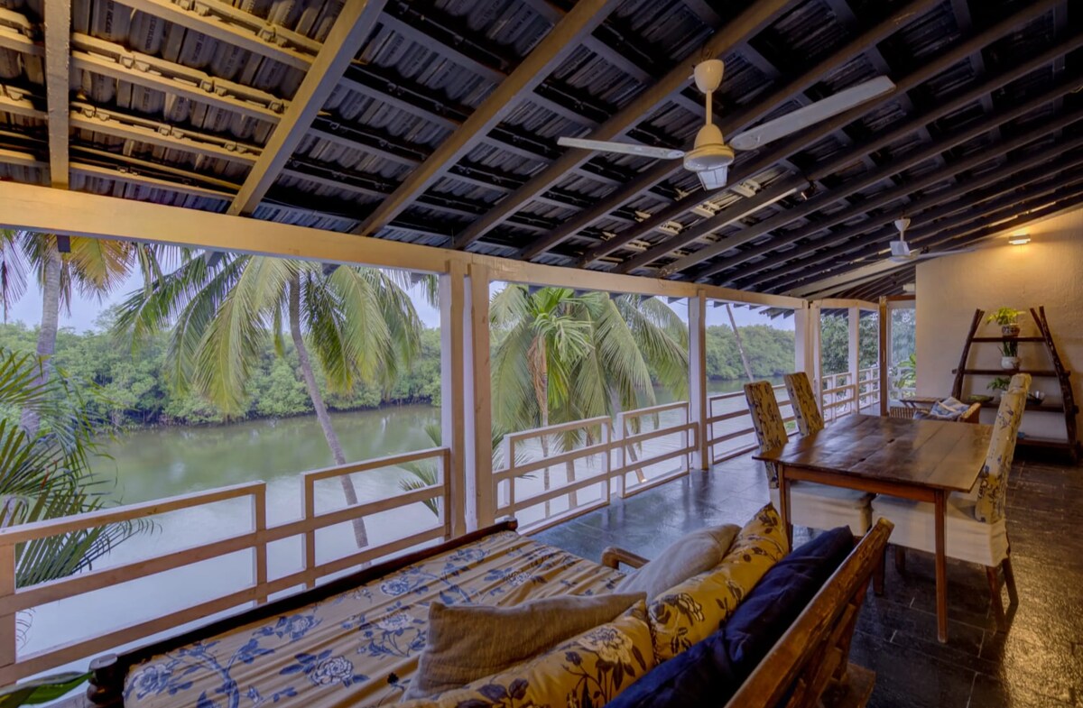 A spacious outdoor seating area is featured, furnished with a long wooden dining table and comfortable seating. Lush palm trees are visible through large open windows, providing a serene view of the river. A ceiling fan hangs above, enhancing airflow.