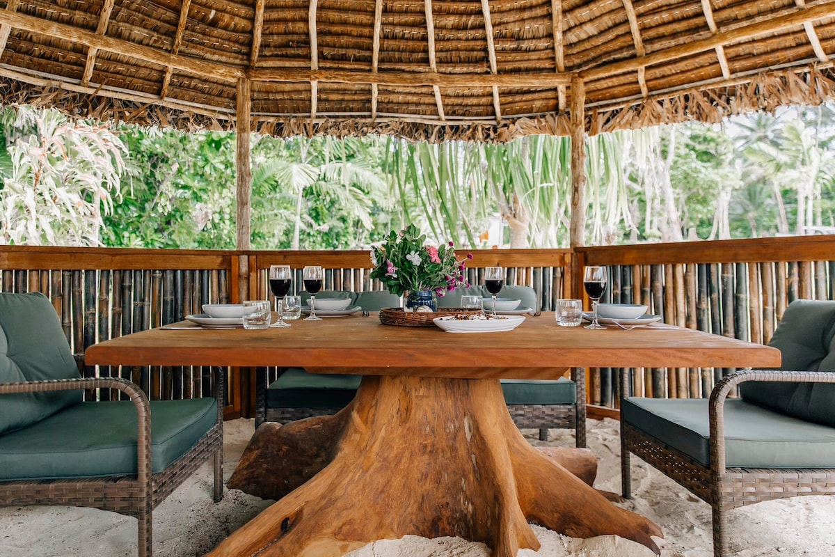 An outdoor dining area is surrounded by a thatched roof, with a large wooden table at its center. Comfortable chairs are positioned around the table, set with plates and glassware. Lush greenery is visible in the background, enhancing the natural ambiance.